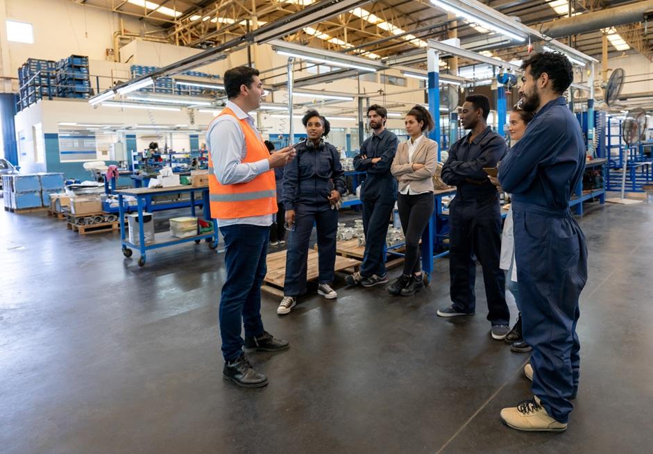 A team of employees pay attention to a manager speaking on the shop floor of a manufacturing facility.