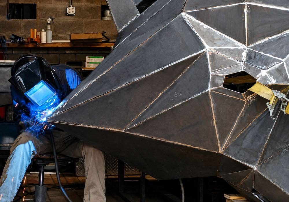 A welder fills a joint on a large raccoon head sculpture.