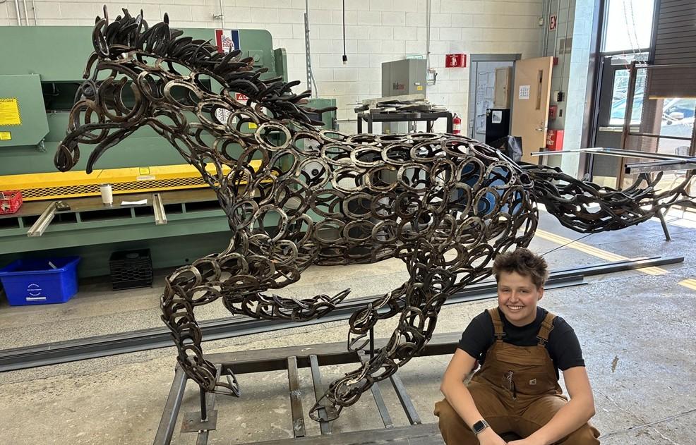 A stuent sits at the base of her metal sculpture of a running horse made from horse shoes..